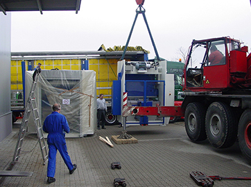 Ein schwerer Kran hebt eine große Maschine vom Transport-LKW, während Arbeiter den Vorgang überwachen.