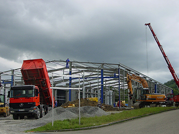 Eine Baustelle mit mehreren Baufahrzeugen, darunter ein Kran und ein Kipplaster, beim Aufbau einer industriellen Halle.