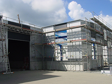Ein Industriebau in der Bauphase mit eingerüsteten Wänden und offenen Halleneingängen.